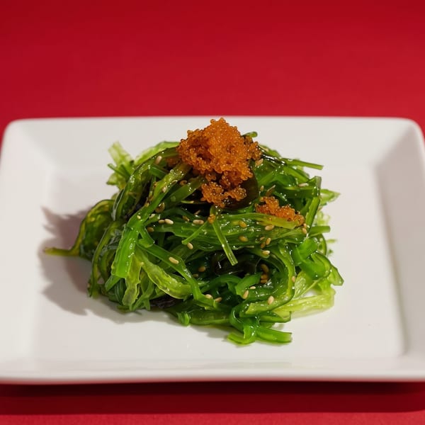 Bright green seaweed salad with orange tobiko and sesame seeds on a white rectangular plate.