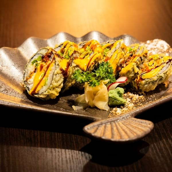 Crispy tempura sushi rolls drizzled with yellow and brown sauces, served on a decorative plate.