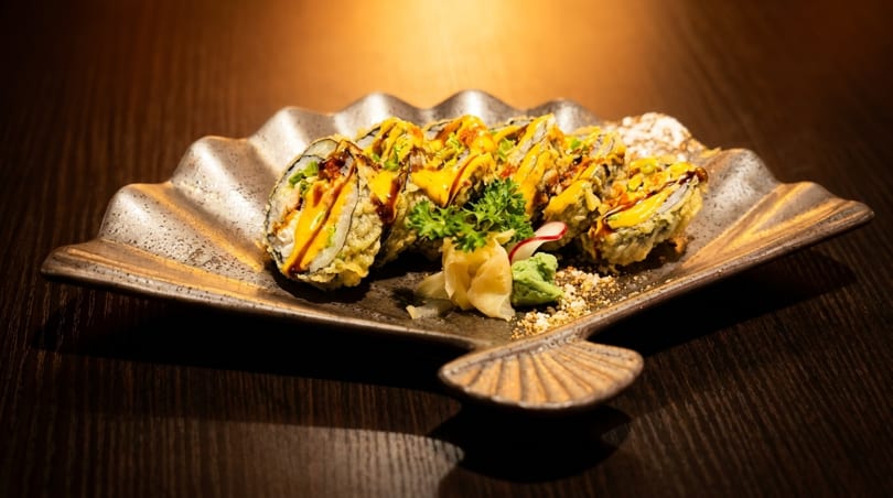 Crispy tempura sushi rolls drizzled with yellow and brown sauces, served on a decorative plate.