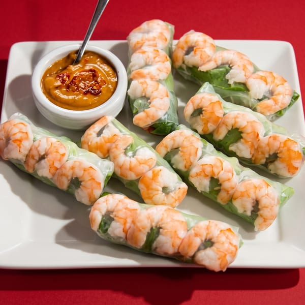 Shrimp spring rolls with peanut dipping sauce on a white rectangular plate against a red background.