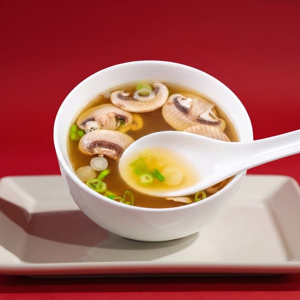 Mushroom soup with green onions in a white bowl and spoon on a white plate against a red background.