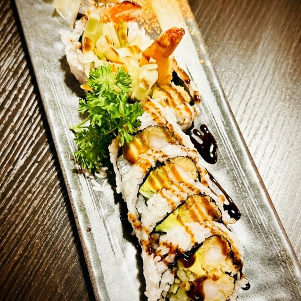 Sushi rolls with shrimp tempura, avocado, and sauce, garnished with parsley on a dark plate.