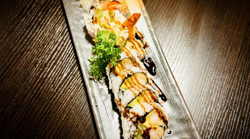 Sushi rolls with shrimp tempura, avocado, and sauce, garnished with parsley on a dark plate.