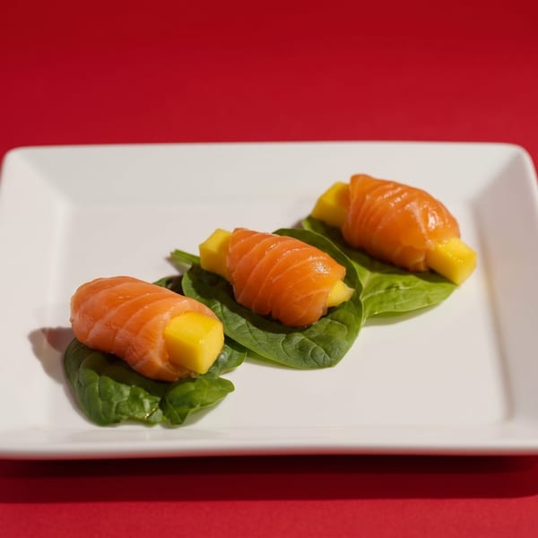 Three salmon and mango rolls on spinach leaves, arranged on a white rectangular plate.