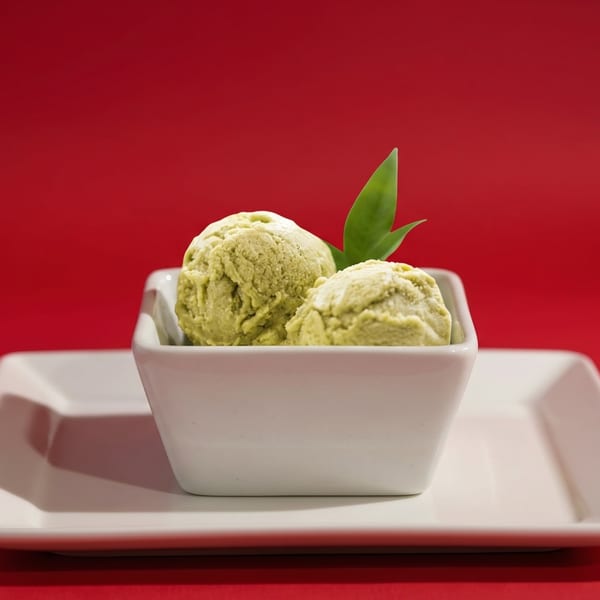 Two scoops of green tea ice cream with a green leaf garnish in a white bowl on a red background.
