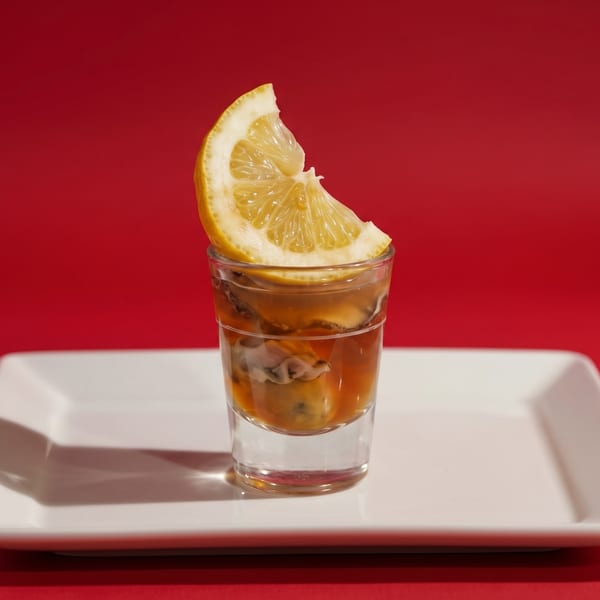 Oysters in a shot glass with a lemon slice, on a white rectangular plate against a red background.